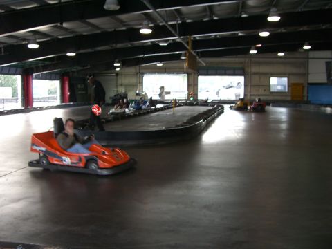 1996 Ford Probe - Go-karts race on an indoor track with spectators in the background.