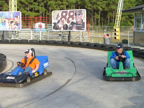 1996 Ford Probe - Two men race go-karts on an outdoor track, with a large billboard race car drivers in the background.