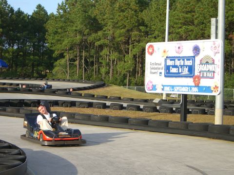 1996 Ford Probe - A person drives a go-kart on a track, with a large sign for Broadway at the Beach visible in the background.