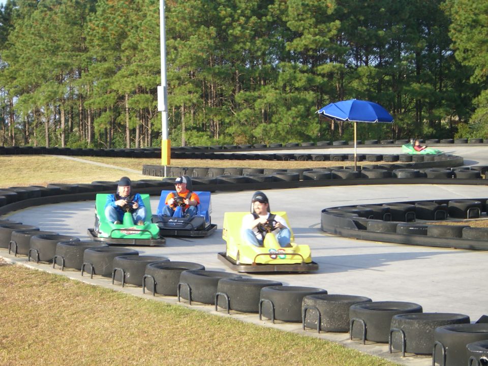 Three go-karts with drivers on a paved race track with tire barriers and trees in the background