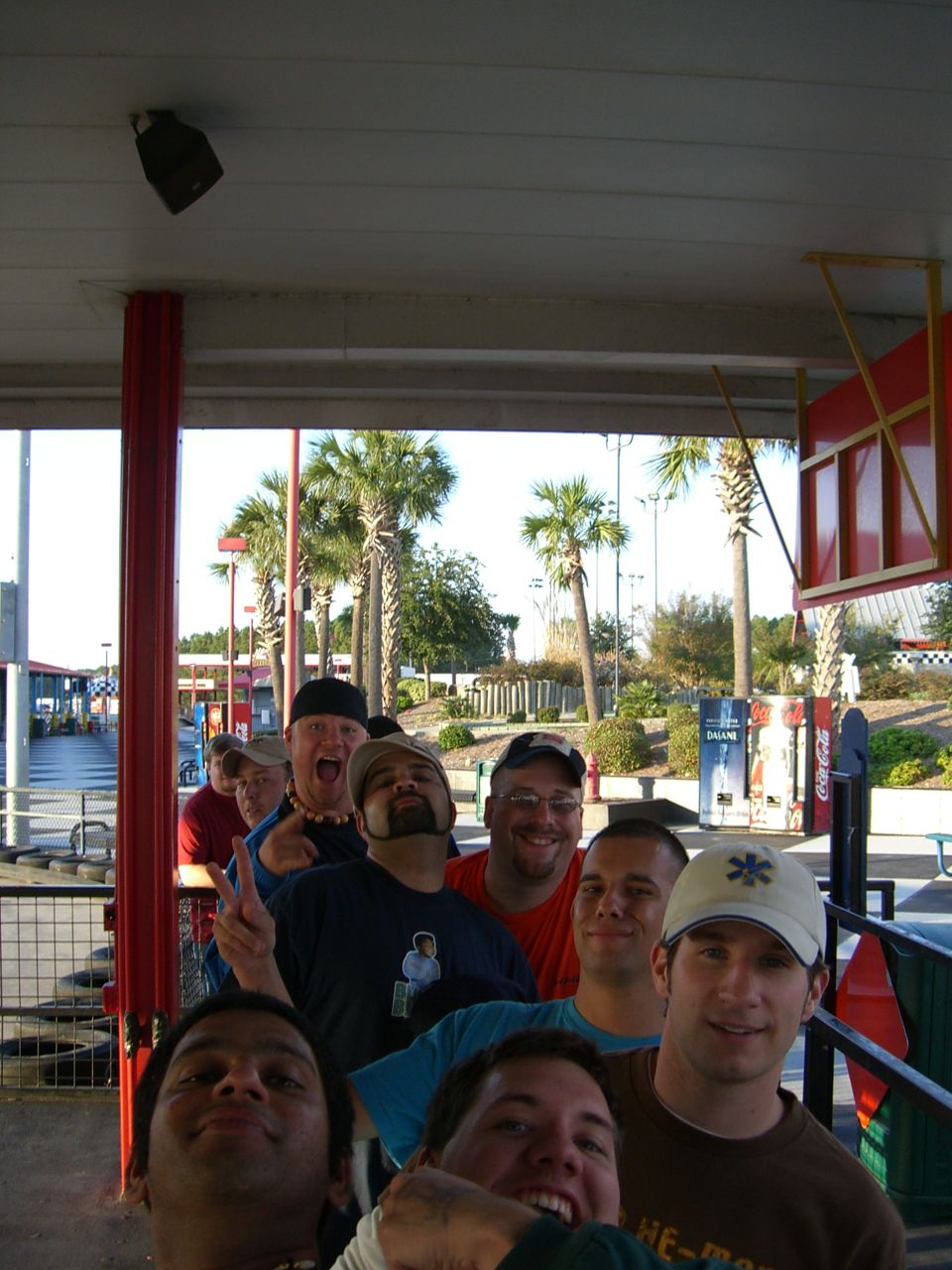 Group of men in line, one making peace sign, palm trees and outdoor structures in background