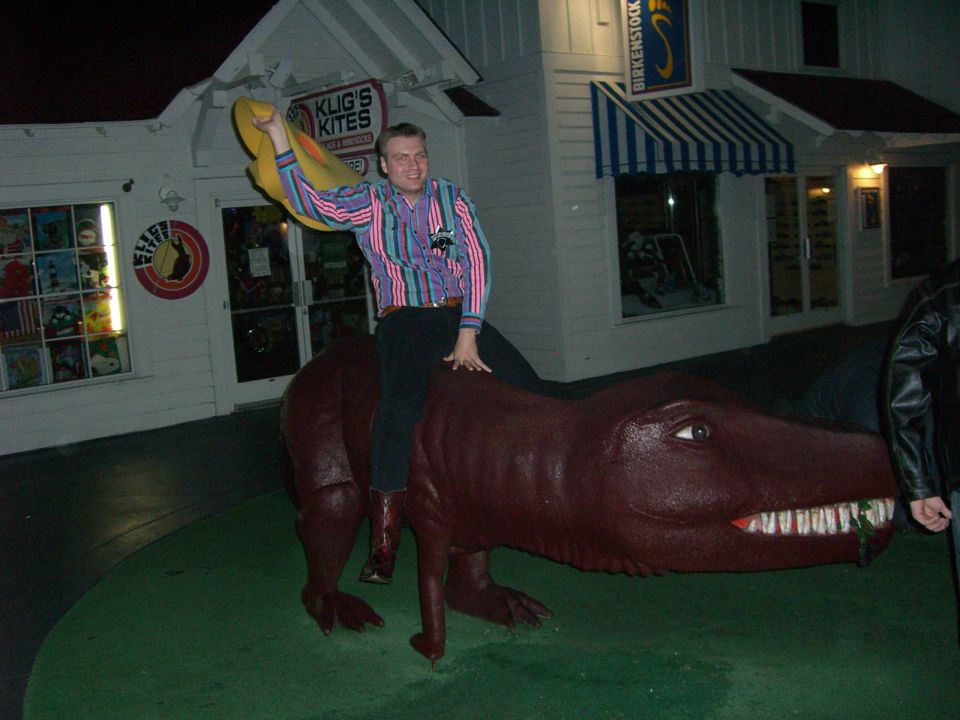 Man in a striped shirt rides a maroon dinosaur statue in front of Klug's Kites store