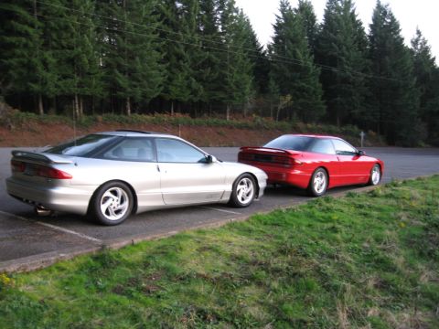 1996 Ford Probe - Two Ford Probes, one silver and one red, are parked next to each other on a paved area next to a grassy slope and trees.