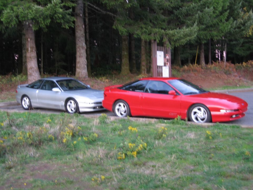 Two 1996 Ford Probes parked side-by-side on a grassy shoulder next to trees