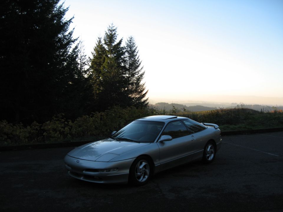 Silver coupe parked on asphalt with trees and hills in background