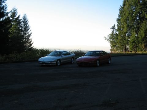 1996 Ford Probe - Two Ford Probe cars are parked side-by-side on a paved area, with a backdrop of trees and a distant hazy landscape.