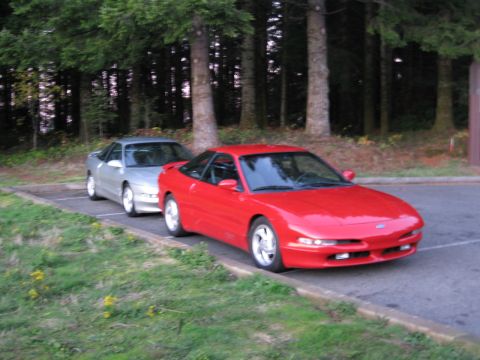 1996 Ford Probe - Two Ford Probes, one red and one silver, are parked side-by-side in a lot with trees in the background.