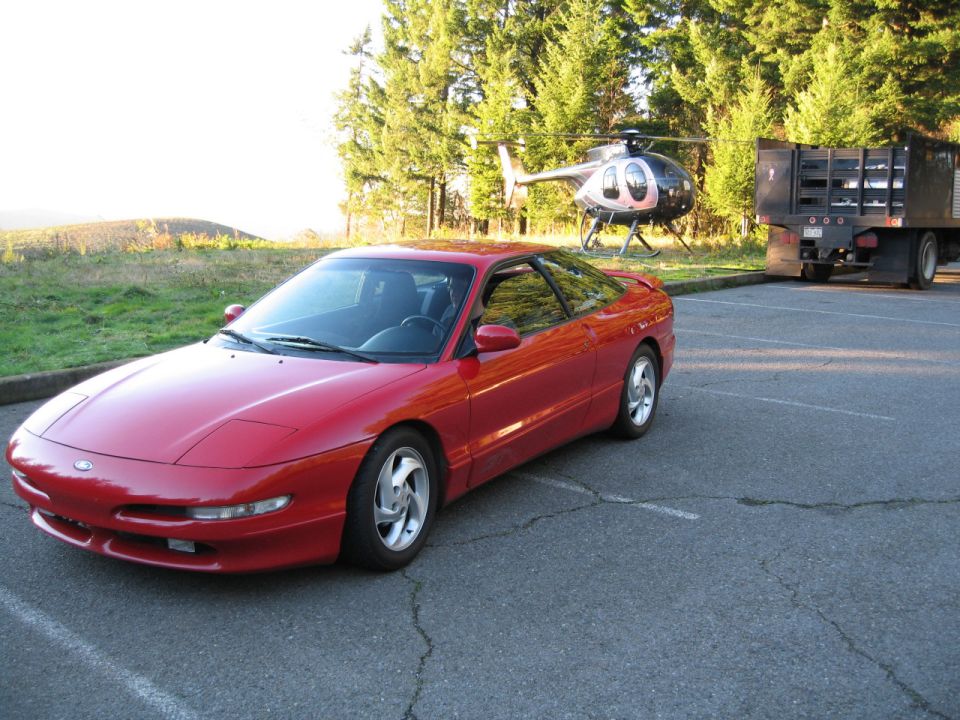 Red Ford Probe coupe front three-quarter view in a parking lot, helicopter behind