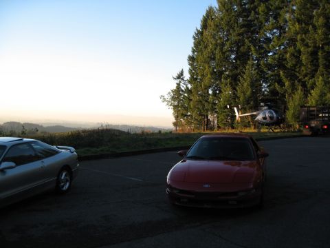 1996 Ford Probe - Two Ford Probes are parked in a lot with a helicopter and a truck in the background, overlooking a misty valley.