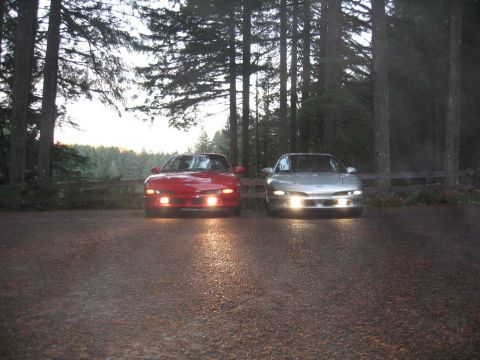 1996 Ford Probe - Two coupes are parked side-by-side in a wooded area with their headlights on.