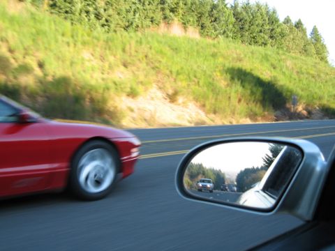 1996 Ford Probe - A red sports car is captured in motion on a two-lane road, with other vehicles visible in the side mirror.