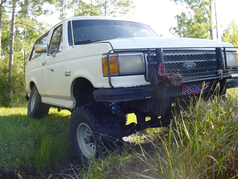 White Ford Bronco with a brush guard and Confederate flag, lifted with large tires, parked in tall grass