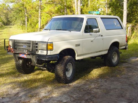 1989 Ford Bronco - This lifted white Ford Bronco sits parked on a dirt road with trees in the background.
