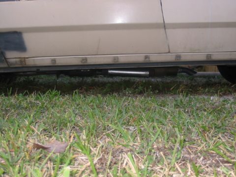 1985 Ford Crown Victoria LTD - The exhaust pipe and lower body panel of a tan vehicle are visible against a grassy background.