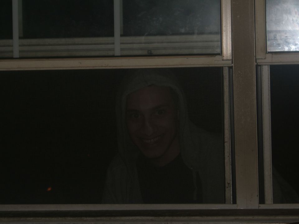 Person with a hood and a wide smile looking through a window at night