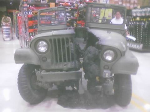 1990 Ford Bronco - Two olive-drab flat-fender military-style Jeeps parked indoors inside a retail store, with model-car shelves and a person in the background.
