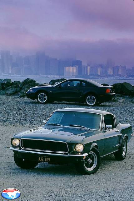 1981 Ford Mustang - Two generations of Ford Mustangs are parked outdoors with a city skyline visible in the distance.