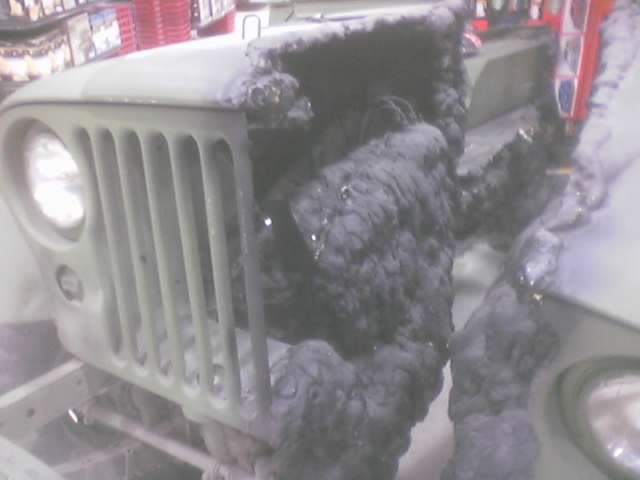 Close-up front of an olive flat-fender Jeep with slotted grille beside a black tire stack