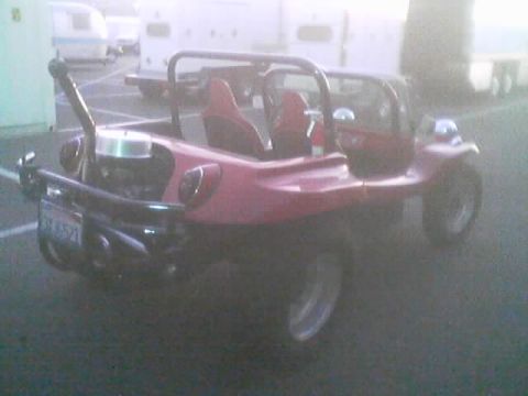 1990 Ford Bronco - pink-red dune buggy with a roll cage and striped seats parked in a misty lot beside white trailers, showing a rear license plate.