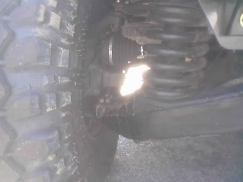1990 Ford Bronco - Close underside view of an off-road tire, coil spring and CV-style boot on a vehicle's front suspension, with bright sunlight glare.