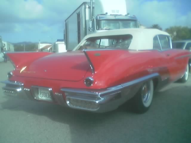 Red convertible with white top and tail fins, license plate 2SOV36. Truck behind it