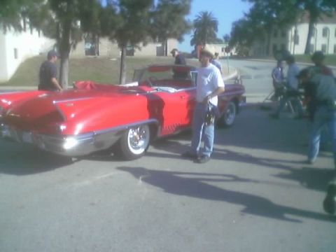 1968 Ford Mustang - A red convertible with prominent fins and a chrome bumper is parked on a street, with a man standing next to it.