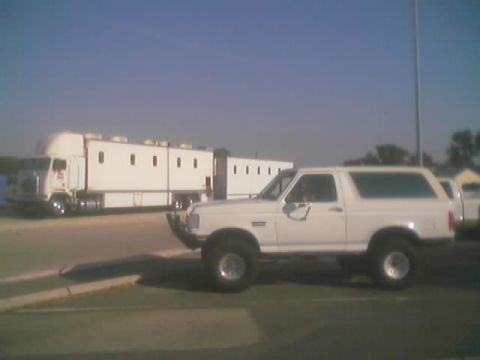1990 Ford Bronco - white lifted two-door Ford Bronco-style SUV parked in a lot next to a white semi truck and box trailer under a clear sky.