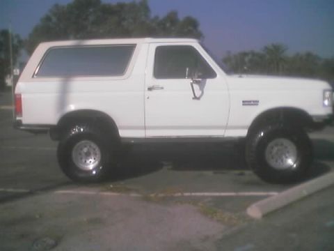1990 Ford Bronco - white lifted two-door Ford Bronco-style SUV with chrome wheels parked in an empty paved lot under a blue sky.