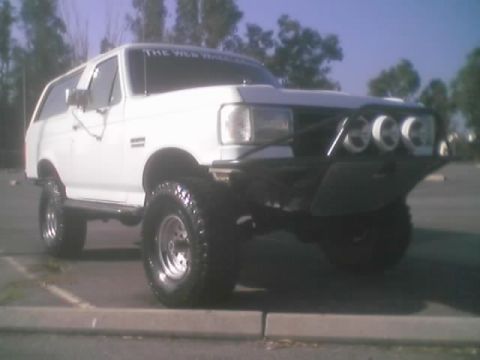 1990 Ford Bronco - red dune buggy with a roll cage and striped bucket seats parked head-on in front of a building, with WINNER signage to the right.