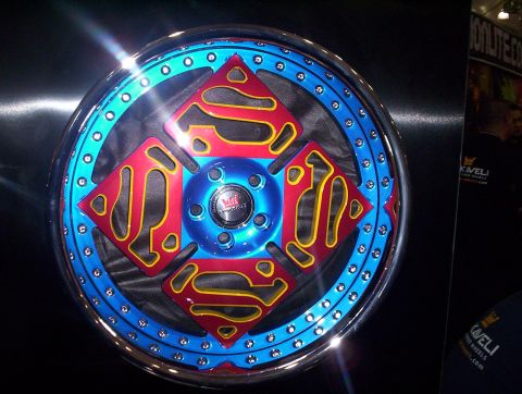 1998 Ford Crown Victoria - custom alloy wheel with a blue rim and red-and-yellow Superman-style center design displayed on a black panel at a show.