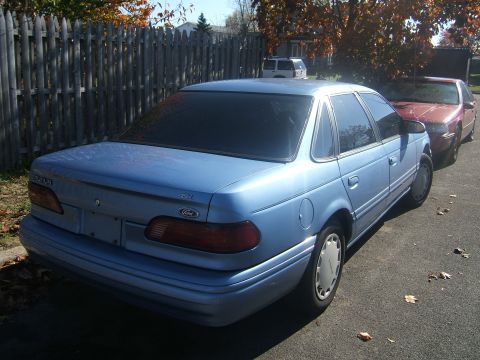 1994 Ford Taurus - A blue sedan is parked on the street next to another vehicle, with a wooden fence in the background.