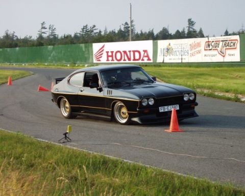 1981 Ford Mustang - A black coupe with gold accents drives on a race track with orange cones marking the course.