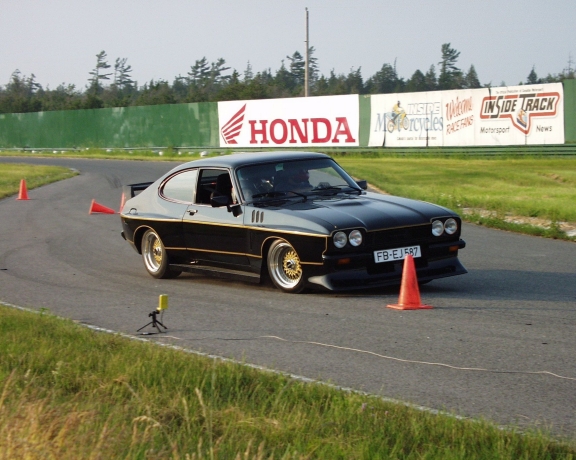 Black coupe with gold trim on race track, orange cones visible
