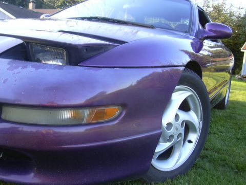 1997 Ford Probe - Detail of a purple coupe's front corner, showing the headlight, amber turn signal and a white five-spoke alloy wheel on grass.