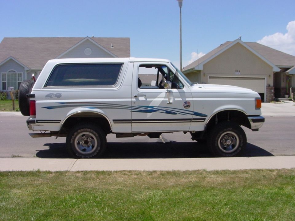 White Ford Bronco SUV with lift, all-terrain tires, and blue stripe graphics, parked curbside, side view