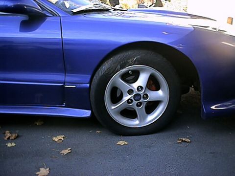 1993 Ford Probe - A close-up view of the front wheel and fender of a blue 1993 Ford Probe, with fallen leaves scattered on the ground.