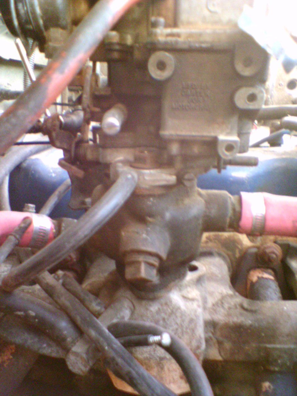 Close-up of a carburetor and adapter spacer stacked on a weathered intake manifold with linkage and hoses