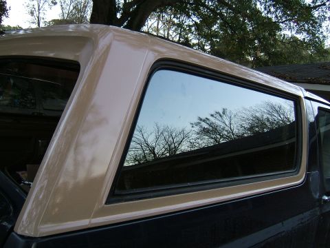 1988 Ford Bronco - The rear side window and tan top of a Ford Bronco are shown, with reflections of trees in the glass.