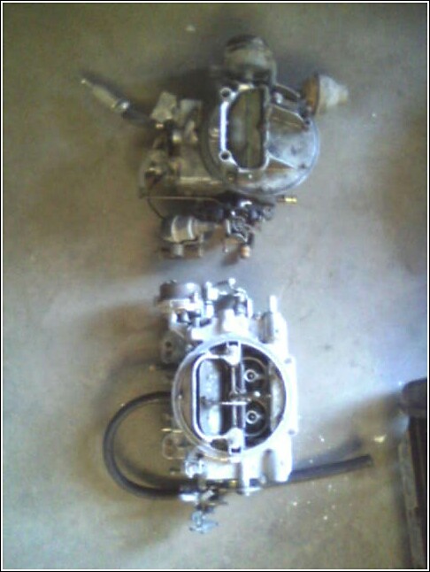 Two carburetors on a concrete floor. One is larger and older looking than the other