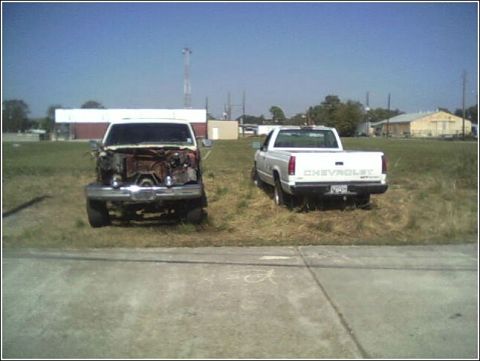 1980 Ford Bronco - Two trucks are parked in a field, one appearing to be stripped for parts or damaged, while the other is a white Chevrolet pickup.
