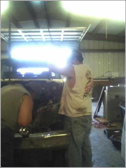 1980 Ford Bronco - Mechanics work on an engine bay inside a garage, with a bright light illuminating the scene.