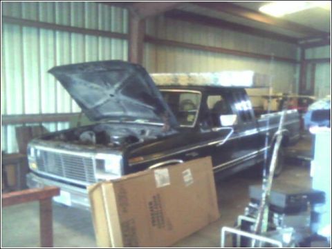1980 Ford Bronco - black square-body Ford pickup with its hood raised inside a metal-sided pole barn, surrounded by boxes and shop equipment.