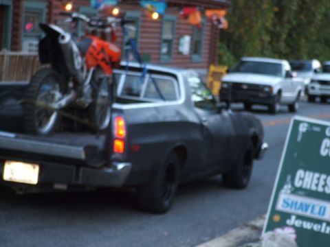 1966 Ford Ranchero - A dark-colored pickup truck transports a dirt bike, passing a roadside sign advertising food items.