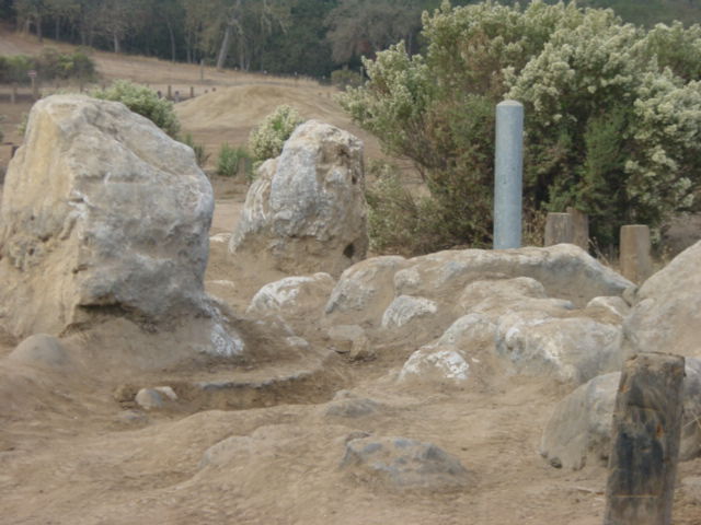 Rocky, sandy landscape with scattered boulders and dry brush under a hazy sky. A gray cylindrical marker stands among the rocks