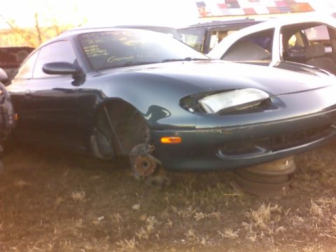 1996 Ford Probe - A dark green coupe, possibly a Mazda MX-5 Miata, is seen in a junkyard with its front wheels removed.
