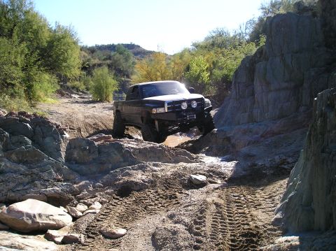 1993 Ford Bronco - A lifted pickup truck maneuvers through a challenging off-road course with rocky terrain and sparse vegetation.