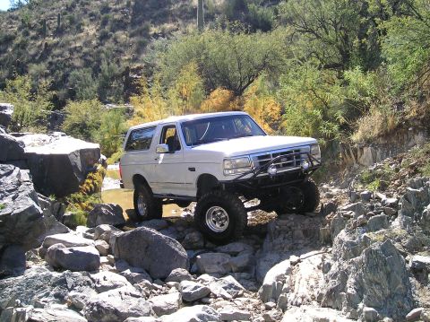 1993 Ford Bronco - White 1993 Ford Bronco tackling a challenging off-road trail with large rocks and water.