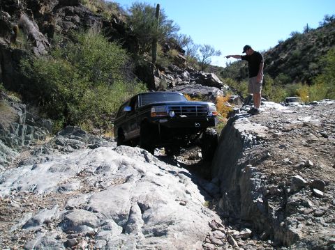 1993 Ford Bronco - A dark blue Ford Bronco is driven off-road through a rocky canyon, with a man observing its path.