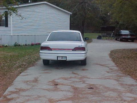 1993 Ford Crown Victoria - The rear of a white sedan is visible, parked on a concrete driveway next to a house.