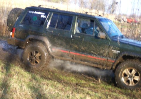 1978 Ford Bronco - Dark green four-door Jeep Cherokee XJ climbing a muddy grass bank, body and mud-terrain tires caked in wet mud. A 'Jeep Jamboree USA' decal…